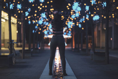 Rear view of woman standing on footpath against illuminated lights at night