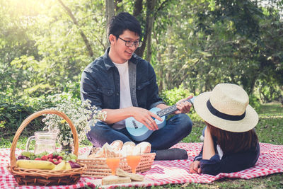 Happy man playing ukulele by girlfriend lying on field during picnic