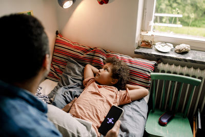 Boy looking at illuminated electric lamp while father holding smart phone on bed at home