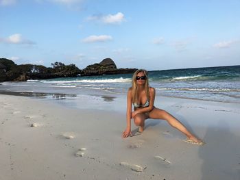 Sensuous woman in bikini crouching at beach against sky