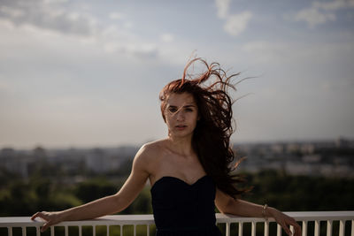 Portrait of young woman with tousled hair standing against sky