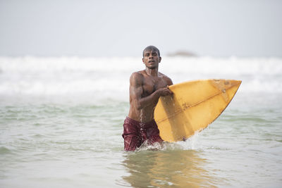 Shirtless man with surfboard wading in sea against sky