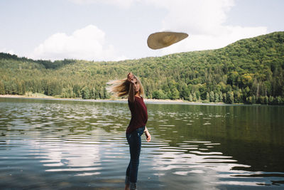Side view full length of young woman throwing hat while standing by lake