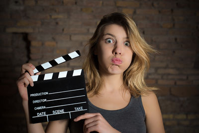 Portrait of young woman making face while holding film slate against brick wall