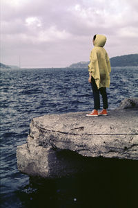 Rear view of woman standing on rock formation by sea against cloudy sky