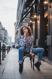 Full length portrait of young woman sitting on chair on sidewalk in city