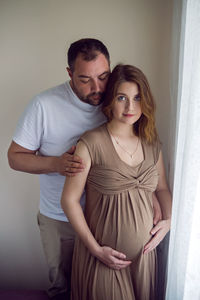 Pregnant girl in a brown dress is standing at the window with her husband in a white t-shirt at home
