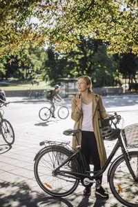 Young female commuter talking on speaker phone while standing with bicycle under tree