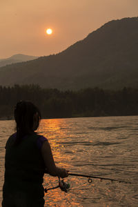 Woman fishing in lake against sky during sunset