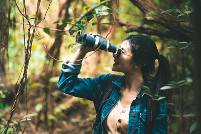 Young woman looking through binoculars while standing in forest