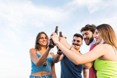 Smiling friends toasting beer bottles while standing against sky