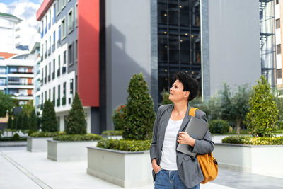 Woman with backpack and laptop walking among modern buildings of business center