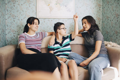 Disabled boy talking to sisters while sitting on sofa against wall at home