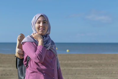 Portrait of young woman standing at beach against sky