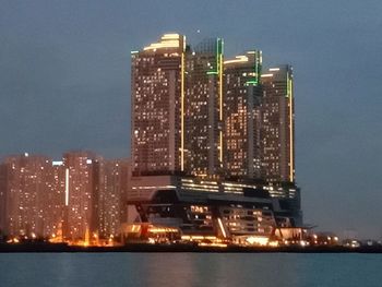 Illuminated modern buildings by sea against sky at night