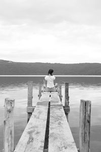 Rear view of woman sitting on pier over lake by mountains against cloudy sky