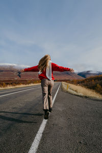 Woman in a red jumper and a scarf running on the road in iceland with arms spread