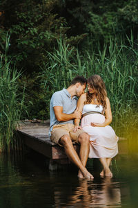 Man kissing pregnant wife while sitting on pier over lake