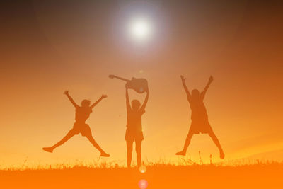 Silhouette siblings with guitar jumping on field against sky during sunset