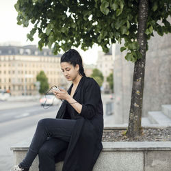 Young woman using smart phone while sitting on retaining wall in city