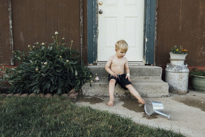 Boy looking at watering can while sitting on steps by door in backyard