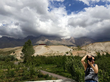 Woman photographing with digital camera on mountain against cloudy sky