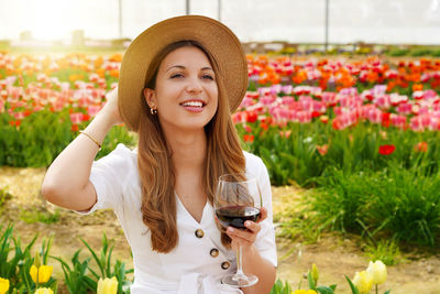 Woman drinking glass red wine over flowered background looking to the side with happy face smiling