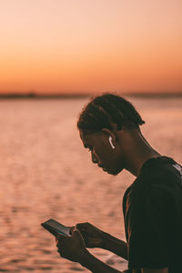 Side view of young man sitting at ocean using mobile phone against clear sky