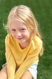 Pretty blonde girl yellow t shirt sitting on green grass on field. top view.