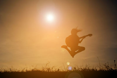 Silhouette girl jumping while playing guitar against sky during sunset