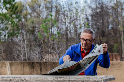 Senior man with glasses reading newspaper at park, front view