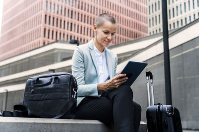 Smiling businesswoman using tablet pc sitting with luggage in front of building