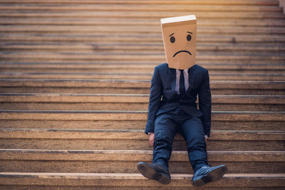 Businessman wearing sad paper mask while sitting on steps in city