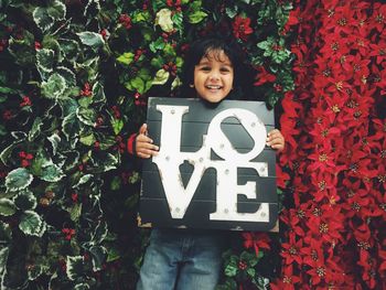 Portrait of happy boy holding love text standing against artificial plants