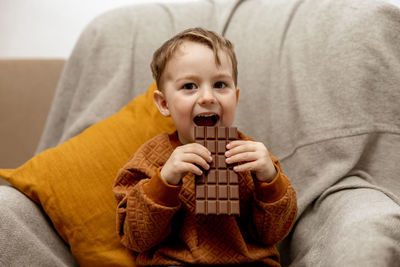 Little adorable boy sitting on the couch at home and eating chocolate bar. child and sweets