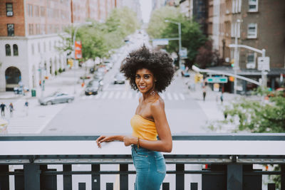 Side view portrait of confident young woman with afro hairstyle standing on bridge against city street