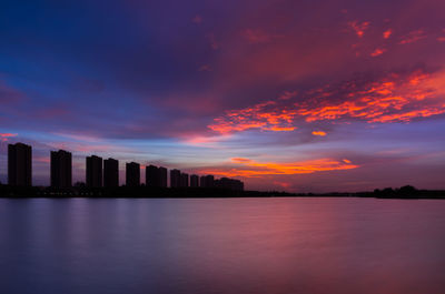 Silhouette buildings by sea against sky during sunset