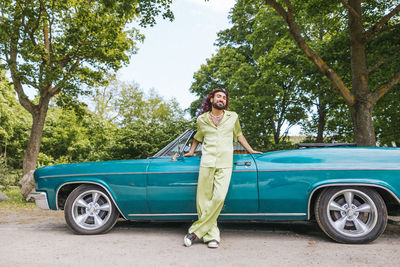Portrait of young man with legs crossed at ankle standing near vintage car on road in park