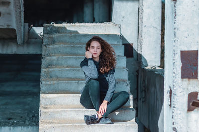 Charming girl with curly hair is sitting on stairs. crossed feet, hand near face.