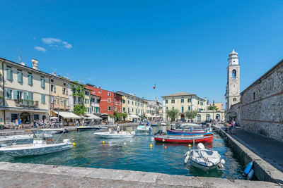 Sailboats moored on canal by buildings against blue sky