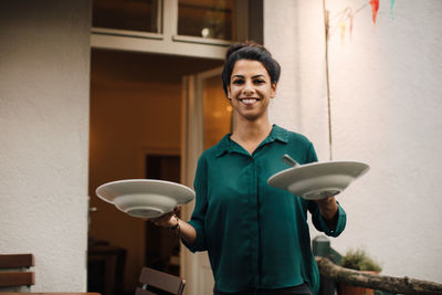 Portrait of smiling young woman holding plates while standing in balcony