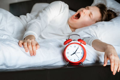 Sleepy millennial woman waking up in bed yawning rubbing eyes turning off alarm clock in bedroom