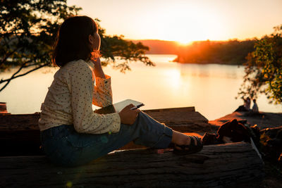 Woman sitting on book by lake against sky during sunset