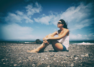 Thoughtful mid adult woman looking away while sitting on shore against cloudy sky