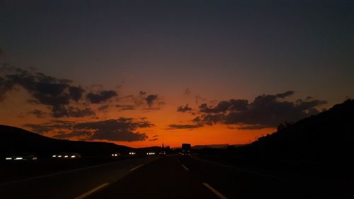 Road by silhouette city against sky during sunset