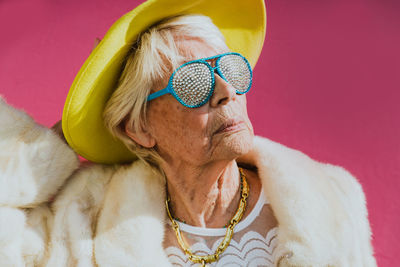 Close-up of senior woman wearing hat and sunglasses against pink background