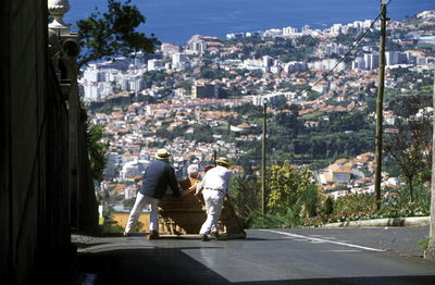 Rear view of men pushing sledge with people on street against cityscape