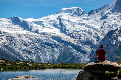 Rear view of man sitting on rock by lake against snowcapped mountains