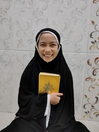 Portrait of smiling girl in hijab sitting against wall at home