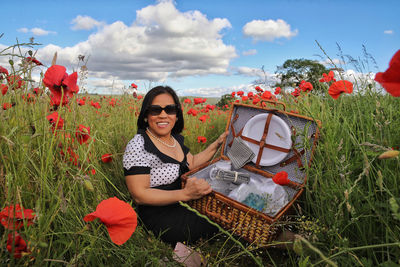 Portrait of happy woman holding picnic basket while sitting amidst red poppy flowers